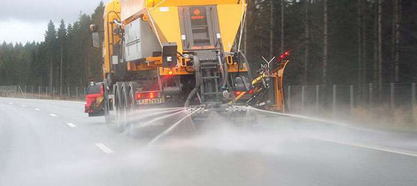 Gul lastbil från Reál Marine sprider vätska på landsväg, vinterväghållning, skog i bakgrunden.